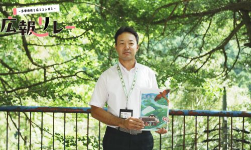 白髭神社境内の木々を背に、奥多摩町 町制70周年記念紙を持った広報担当の森田さんの写真。